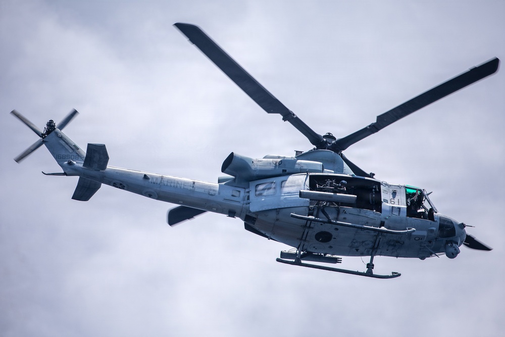 A U.S. Marines military helicopter is photographed in flight against an overcast sky. The aircraft appears to be a Bell UH-1Y Venom or similar variant, displaying 'MARINES' markings and the number '61' on its fuselage. The low-angle shot captures the underside and side profile of the helicopter in detail.