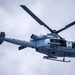 A U.S. Marines military helicopter is photographed in flight against an overcast sky. The aircraft appears to be a Bell UH-1Y Venom or similar variant, displaying 'MARINES' markings and the number '61' on its fuselage. The low-angle shot captures the underside and side profile of the helicopter in detail.