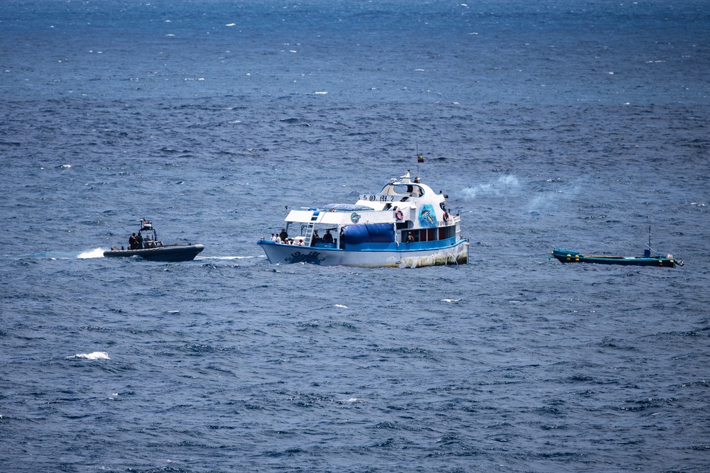 The image shows three vessels on open ocean waters, with a larger blue-and-white passenger or tour boat at center, flanked by two smaller rigid inflatable boats (RIBs). The scene appears to depict a maritime interdiction or law enforcement operation, with what appears to be smoke or exhaust visible near the center vessel. The smaller vessels appear to be tactical or patrol craft approaching or surrounding the larger boat.