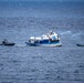 The image shows three vessels on open ocean waters, with a larger blue-and-white passenger or tour boat at center, flanked by two smaller rigid inflatable boats (RIBs). The scene appears to depict a maritime interdiction or law enforcement operation, with what appears to be smoke or exhaust visible near the center vessel. The smaller vessels appear to be tactical or patrol craft approaching or surrounding the larger boat.