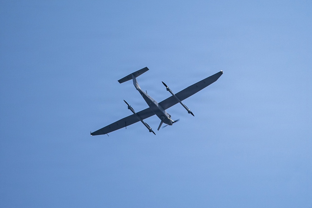 An aircraft is photographed from below against a clear blue sky. The aircraft appears to be a twin-engine turboprop plane with a high-wing configuration, viewed from a low angle showing its underside. Both propellers appear to be spinning, indicating the aircraft is in active flight.