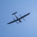 An aircraft is photographed from below against a clear blue sky. The aircraft appears to be a twin-engine turboprop plane with a high-wing configuration, viewed from a low angle showing its underside. Both propellers appear to be spinning, indicating the aircraft is in active flight.