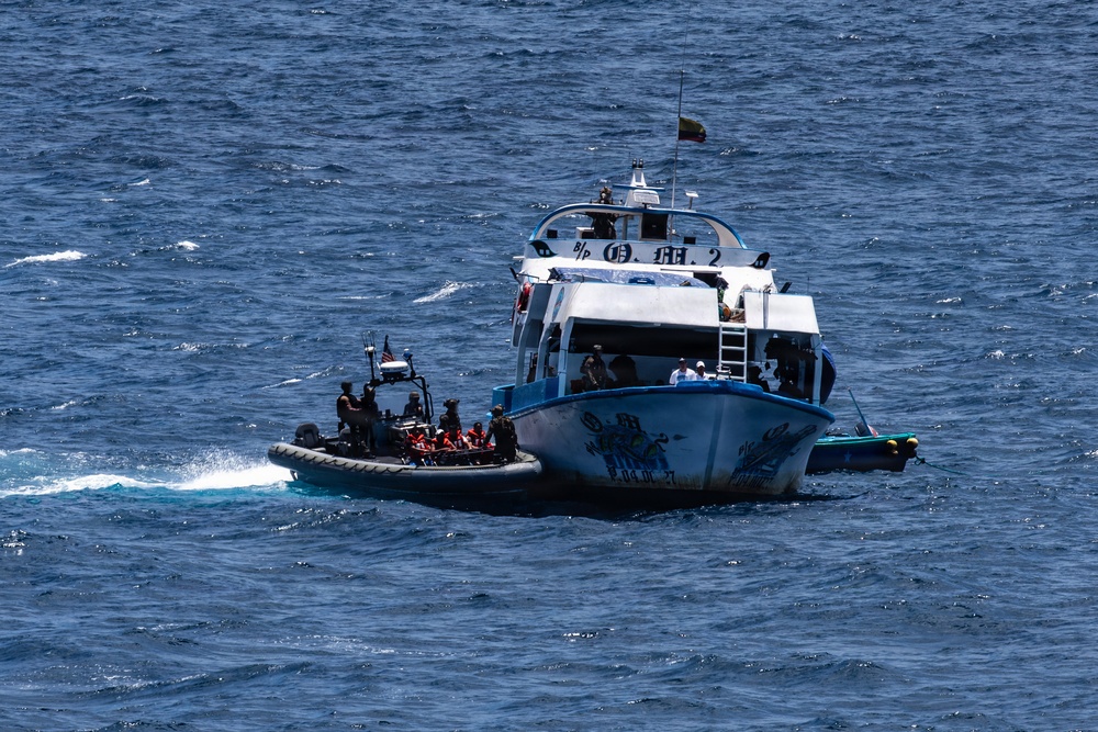A rigid inflatable boat (RIB) with armed personnel in dark tactical gear is approaching or intercepting a larger civilian-style vessel on open blue ocean waters. The larger vessel appears to be a patrol or ferry-type boat flying what appears to be a yellow, red, and green flag. This appears to be a maritime interdiction or law enforcement operation.