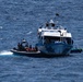 A rigid inflatable boat (RIB) with armed personnel in dark tactical gear is approaching or intercepting a larger civilian-style vessel on open blue ocean waters. The larger vessel appears to be a patrol or ferry-type boat flying what appears to be a yellow, red, and green flag. This appears to be a maritime interdiction or law enforcement operation.