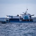 A white and blue fishing/charter boat named 'Manta' with registration number P.04 00727 is navigating choppy open ocean waters. The vessel features decorative artwork of a marlin/swordfish on its hull and appears to be a mid-sized sport fishing or dive charter boat. The sea conditions appear moderately rough with visible wave action.