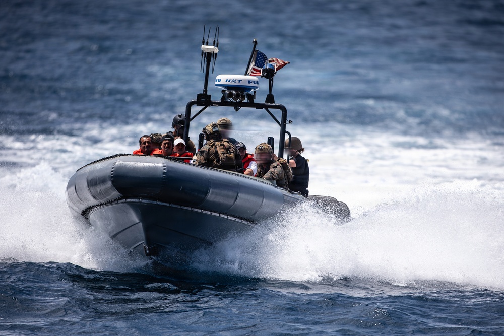 A rigid-hull inflatable boat (RHIB) is underway at high speed on open ocean water, carrying multiple personnel including individuals in military/tactical gear and others in orange life vests. An American flag is visible flying from the boat's console mast alongside navigation and radar equipment. The boat is generating significant wake and spray, indicating high-speed travel.