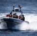 A rigid-hull inflatable boat (RHIB) is underway at high speed on open ocean water, carrying multiple personnel including individuals in military/tactical gear and others in orange life vests. An American flag is visible flying from the boat's console mast alongside navigation and radar equipment. The boat is generating significant wake and spray, indicating high-speed travel.