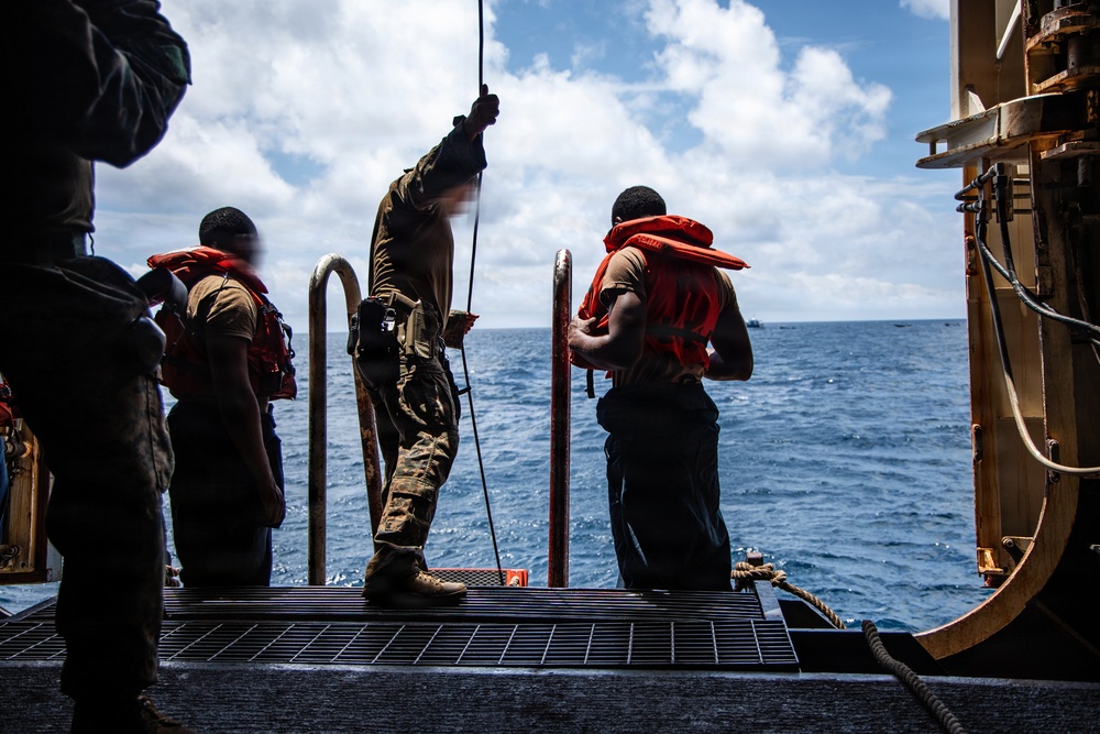 Several military or naval personnel are standing on the deck of a large vessel at the water's edge, wearing orange inflatable life vests. One individual in camouflage uniform is holding a rope or line, while others appear to be preparing for a water-based operation. The scene takes place in open ocean under a partly cloudy sky.