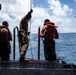 Several military or naval personnel are standing on the deck of a large vessel at the water's edge, wearing orange inflatable life vests. One individual in camouflage uniform is holding a rope or line, while others appear to be preparing for a water-based operation. The scene takes place in open ocean under a partly cloudy sky.