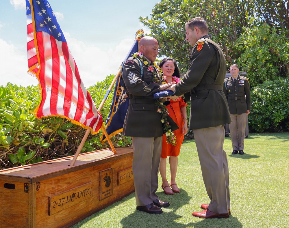 A military retirement ceremony is taking place outdoors, likely in Hawaii based on the tropical vegetation and floral leis worn by participants. A senior non-commissioned officer wearing multiple leis is receiving a folded American flag from a higher-ranking officer, a traditional honor at military retirement ceremonies. A wooden crate marked '2-27 INFANTRY' and 'THE WOLF' is visible in the foreground, likely used to transport unit colors or ceremonial items.