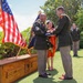 A military retirement ceremony is taking place outdoors, likely in Hawaii based on the tropical vegetation and floral leis worn by participants. A senior non-commissioned officer wearing multiple leis is receiving a folded American flag from a higher-ranking officer, a traditional honor at military retirement ceremonies. A wooden crate marked '2-27 INFANTRY' and 'THE WOLF' is visible in the foreground, likely used to transport unit colors or ceremonial items.