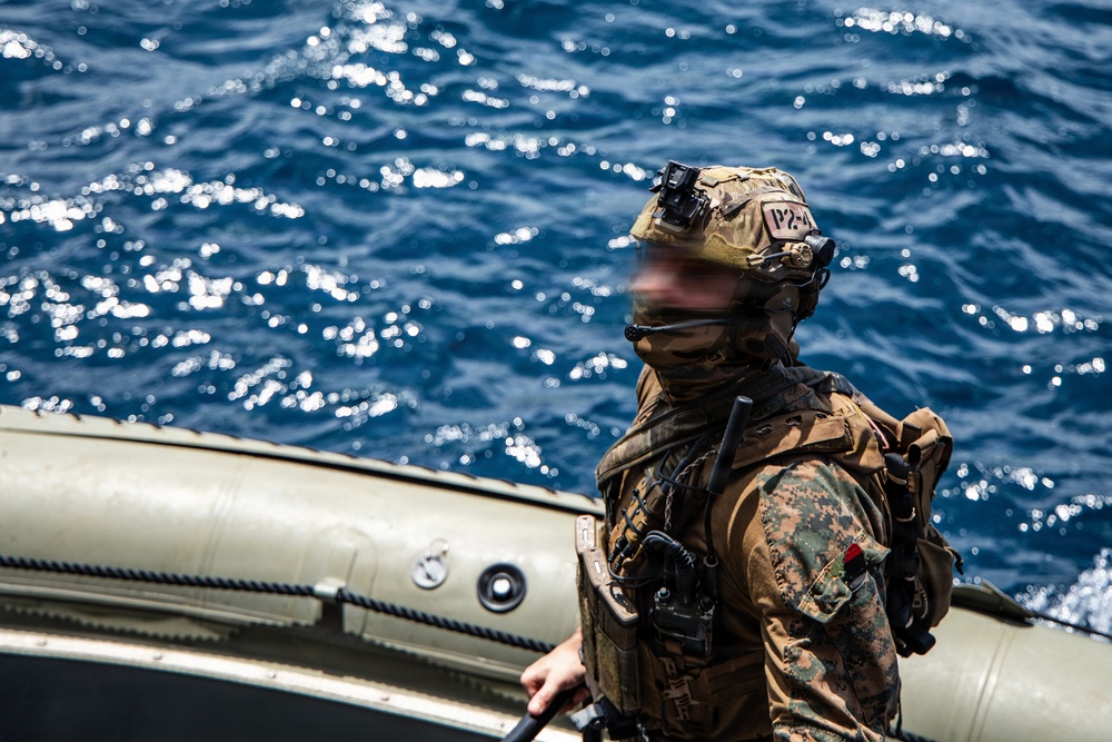 A heavily equipped military operator stands aboard what appears to be a rigid inflatable boat (RIB) on open ocean waters. The individual is wearing full tactical gear including a camouflage uniform, combat helmet with mounted device, face covering, and communications equipment. The face has been digitally blurred, consistent with operational security practices for special operations personnel.