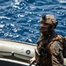 A heavily equipped military operator stands aboard what appears to be a rigid inflatable boat (RIB) on open ocean waters. The individual is wearing full tactical gear including a camouflage uniform, combat helmet with mounted device, face covering, and communications equipment. The face has been digitally blurred, consistent with operational security practices for special operations personnel.
