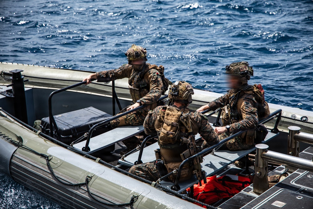 Three military personnel in full combat gear are seated aboard a rigid-hull inflatable boat (RHIB) on open ocean water. The individuals are wearing camouflage uniforms, tactical helmets with mounted equipment, and carrying weapons and gear. One face has been deliberately blurred, suggesting operational security considerations.
