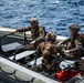 Three military personnel in full combat gear are seated aboard a rigid-hull inflatable boat (RHIB) on open ocean water. The individuals are wearing camouflage uniforms, tactical helmets with mounted equipment, and carrying weapons and gear. One face has been deliberately blurred, suggesting operational security considerations.