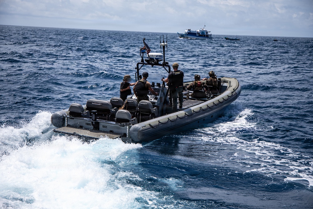 A rigid-hull inflatable boat (RHIB) with hull markings '11MRX 1001' is underway at speed on open ocean, carrying several personnel in tactical gear and body armor. An American flag is visible on a mast-mounted pole, and the crew appears to be conducting a maritime law enforcement or interdiction operation. In the background, a larger blue-and-white vessel and what appears to be a smaller craft are visible on the horizon.