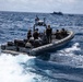 A rigid-hull inflatable boat (RHIB) with hull markings '11MRX 1001' is underway at speed on open ocean, carrying several personnel in tactical gear and body armor. An American flag is visible on a mast-mounted pole, and the crew appears to be conducting a maritime law enforcement or interdiction operation. In the background, a larger blue-and-white vessel and what appears to be a smaller craft are visible on the horizon.