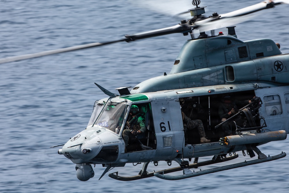 A U.S. military helicopter, identified as aircraft number 61, is captured in flight over open water. The helicopter appears to be a UH-1Y Venom (Huey) variant used by the U.S. Marine Corps, with crew members and door gunners visible with mounted weapons. The image is a close-range action shot showing the aircraft in active operational configuration.