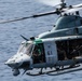 A U.S. military helicopter, identified as aircraft number 61, is captured in flight over open water. The helicopter appears to be a UH-1Y Venom (Huey) variant used by the U.S. Marine Corps, with crew members and door gunners visible with mounted weapons. The image is a close-range action shot showing the aircraft in active operational configuration.