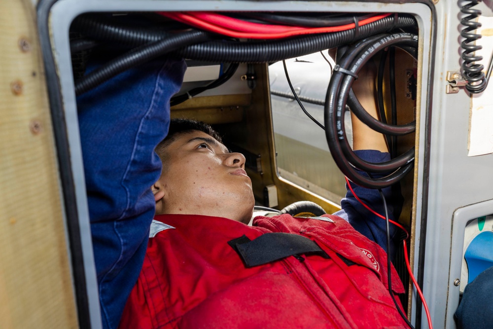A young technician in a red work uniform is lying on his back inside a confined access panel or equipment bay, looking upward while performing maintenance or inspection work. The space contains multiple cables and hoses including red, black, and corrugated conduit lines. The setting appears to be industrial or vehicular equipment maintenance.