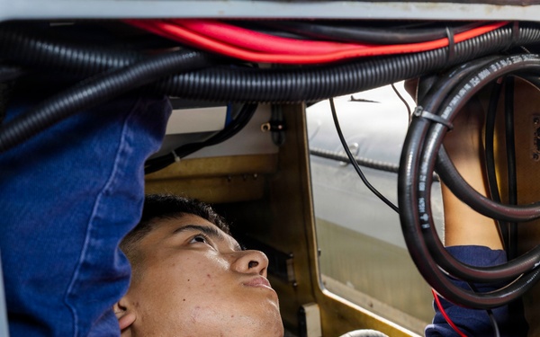 A young technician in a red work uniform is lying on his back inside a confined access panel or equipment bay, looking upward while performing maintenance or inspection work. The space contains multiple cables and hoses including red, black, and corrugated conduit lines. The setting appears to be industrial or vehicular equipment maintenance.