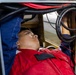 A young technician in a red work uniform is lying on his back inside a confined access panel or equipment bay, looking upward while performing maintenance or inspection work. The space contains multiple cables and hoses including red, black, and corrugated conduit lines. The setting appears to be industrial or vehicular equipment maintenance.