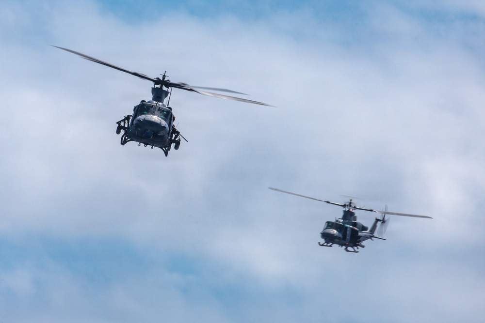 Two military helicopters are flying in formation against a partly cloudy blue sky. The foreground helicopter is larger and closer to the camera, showing significant detail, while a second helicopter flies in the background at a lower altitude. Both aircraft appear to be dark-colored military variants with rotor blades in motion.