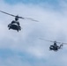 Two military helicopters are flying in formation against a partly cloudy blue sky. The foreground helicopter is larger and closer to the camera, showing significant detail, while a second helicopter flies in the background at a lower altitude. Both aircraft appear to be dark-colored military variants with rotor blades in motion.