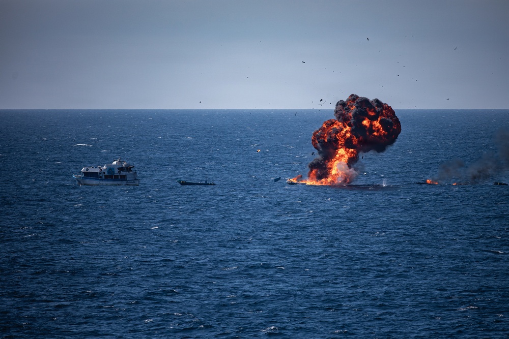 A large explosion with orange flames and black smoke is visible on the open sea, appearing to engulf or destroy a vessel. At least one additional white boat is visible nearby on the water. Debris and what appear to be birds are scattered in the sky around the explosion.