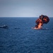 A large explosion with orange flames and black smoke is visible on the open sea, appearing to engulf or destroy a vessel. At least one additional white boat is visible nearby on the water. Debris and what appear to be birds are scattered in the sky around the explosion.