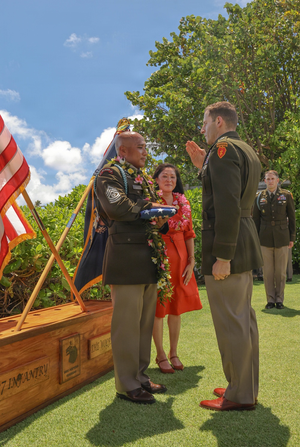An outdoor U.S. Army military ceremony, likely a retirement or change of command, taking place on a grass lawn in what appears to be Hawaii. A senior non-commissioned officer wearing traditional Hawaiian lei garlands holds a folded American flag while facing an officer who is rendering a salute. A woman in a red dress, also wearing leis, stands between them alongside other uniformed soldiers in the background.