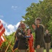 An outdoor U.S. Army military ceremony, likely a retirement or change of command, taking place on a grass lawn in what appears to be Hawaii. A senior non-commissioned officer wearing traditional Hawaiian lei garlands holds a folded American flag while facing an officer who is rendering a salute. A woman in a red dress, also wearing leis, stands between them alongside other uniformed soldiers in the background.