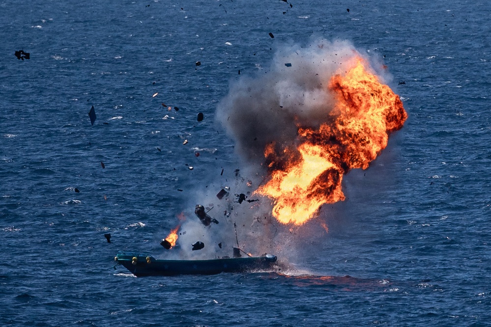 A small watercraft on open ocean is engulfed in a large explosion with intense orange flames and dark smoke rising upward. Debris is visibly scattered in multiple directions from the blast epicenter. The vessel appears to be a small boat, partially intact at the waterline while the upper portion is being destroyed by the explosion.