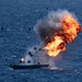 A small watercraft on open ocean is engulfed in a large explosion with intense orange flames and dark smoke rising upward. Debris is visibly scattered in multiple directions from the blast epicenter. The vessel appears to be a small boat, partially intact at the waterline while the upper portion is being destroyed by the explosion.