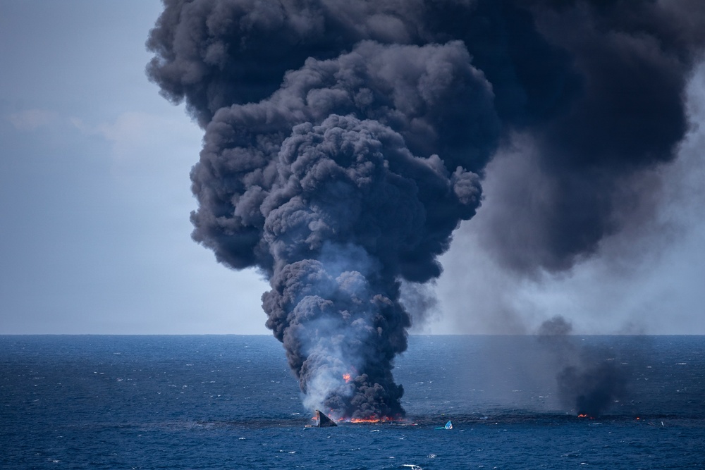 A large fire is burning on the open ocean surface, producing a massive column of dense black smoke rising high into the overcast sky. The fire appears to be consuming a vessel or floating debris, with visible orange flames at the water surface. The scale and color of the smoke column suggests combustion of petroleum-based materials.