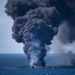A large fire is burning on the open ocean surface, producing a massive column of dense black smoke rising high into the overcast sky. The fire appears to be consuming a vessel or floating debris, with visible orange flames at the water surface. The scale and color of the smoke column suggests combustion of petroleum-based materials.