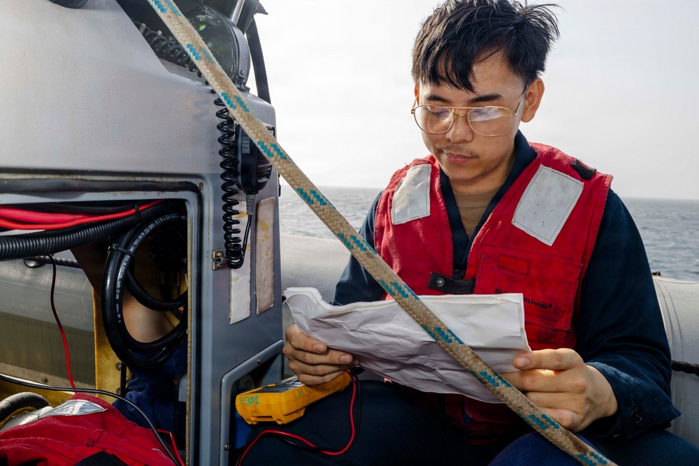 A young man wearing a red life vest and gold-framed glasses is seated on a small watercraft, reviewing a paper document. He is positioned next to an equipment panel containing a radio/communication device, wiring, and what appears to be a yellow multimeter. The setting is on open water under an overcast sky.