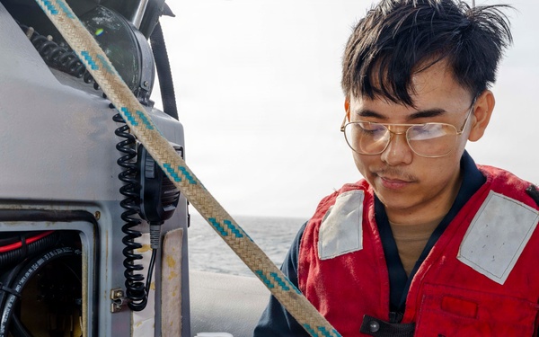 A young man wearing a red life vest and gold-framed glasses is seated on a small watercraft, reviewing a paper document. He is positioned next to an equipment panel containing a radio/communication device, wiring, and what appears to be a yellow multimeter. The setting is on open water under an overcast sky.