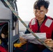 A young man wearing a red life vest and gold-framed glasses is seated on a small watercraft, reviewing a paper document. He is positioned next to an equipment panel containing a radio/communication device, wiring, and what appears to be a yellow multimeter. The setting is on open water under an overcast sky.