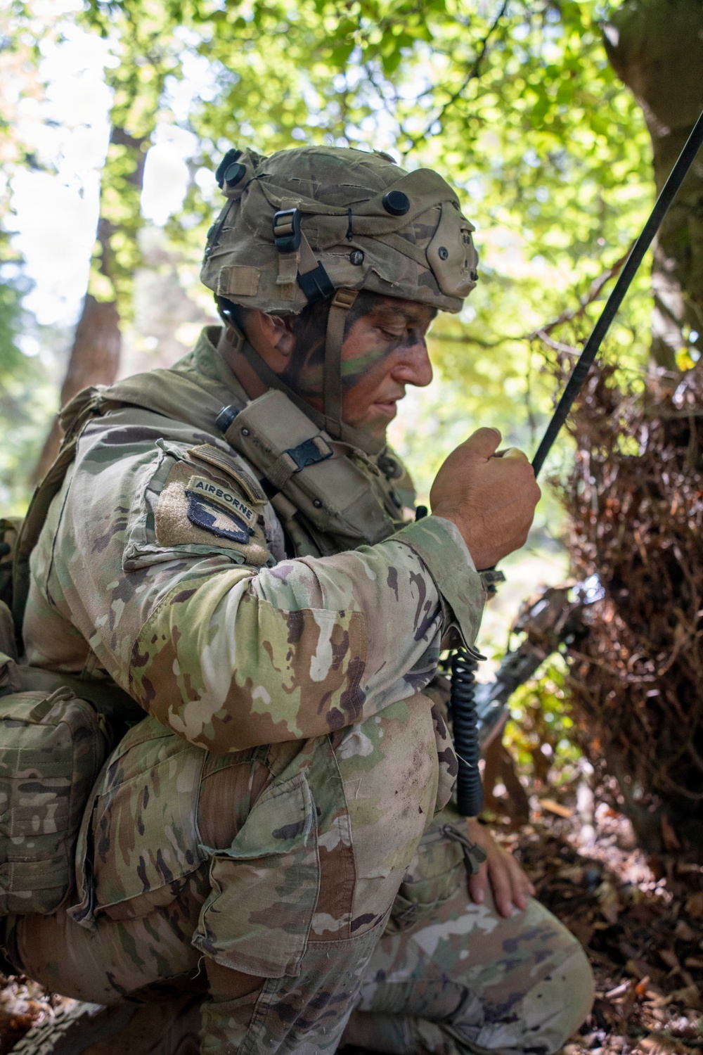 A U.S. Army Airborne soldier in full combat gear is crouched in a wooded area, operating a tactical radio with an extended antenna. The soldier is wearing MultiCam (OCP) camouflage uniform, a combat helmet with accessories, and has face paint applied for concealment. The setting appears to be a field training exercise or operational environment in a forested location.