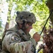 A U.S. Army Airborne soldier in full combat gear is crouched in a wooded area, operating a tactical radio with an extended antenna. The soldier is wearing MultiCam (OCP) camouflage uniform, a combat helmet with accessories, and has face paint applied for concealment. The setting appears to be a field training exercise or operational environment in a forested location.