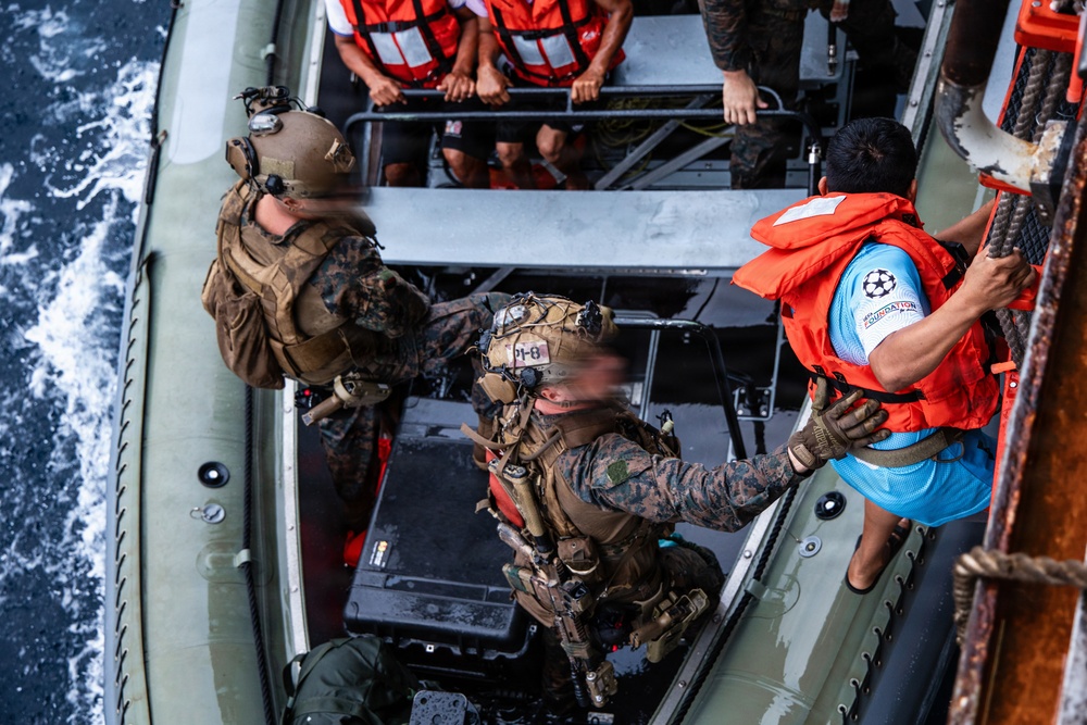 An aerial/overhead view of a military maritime boarding operation showing armed personnel in combat gear on an inflatable rigid-hull boat (RHIB) alongside a larger vessel. Two soldiers in camouflage uniforms and tactical gear are assisting or boarding individuals wearing orange life vests. Multiple civilians in orange life vests are visible seated on the upper deck of the larger vessel.