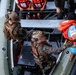 An aerial/overhead view of a military maritime boarding operation showing armed personnel in combat gear on an inflatable rigid-hull boat (RHIB) alongside a larger vessel. Two soldiers in camouflage uniforms and tactical gear are assisting or boarding individuals wearing orange life vests. Multiple civilians in orange life vests are visible seated on the upper deck of the larger vessel.