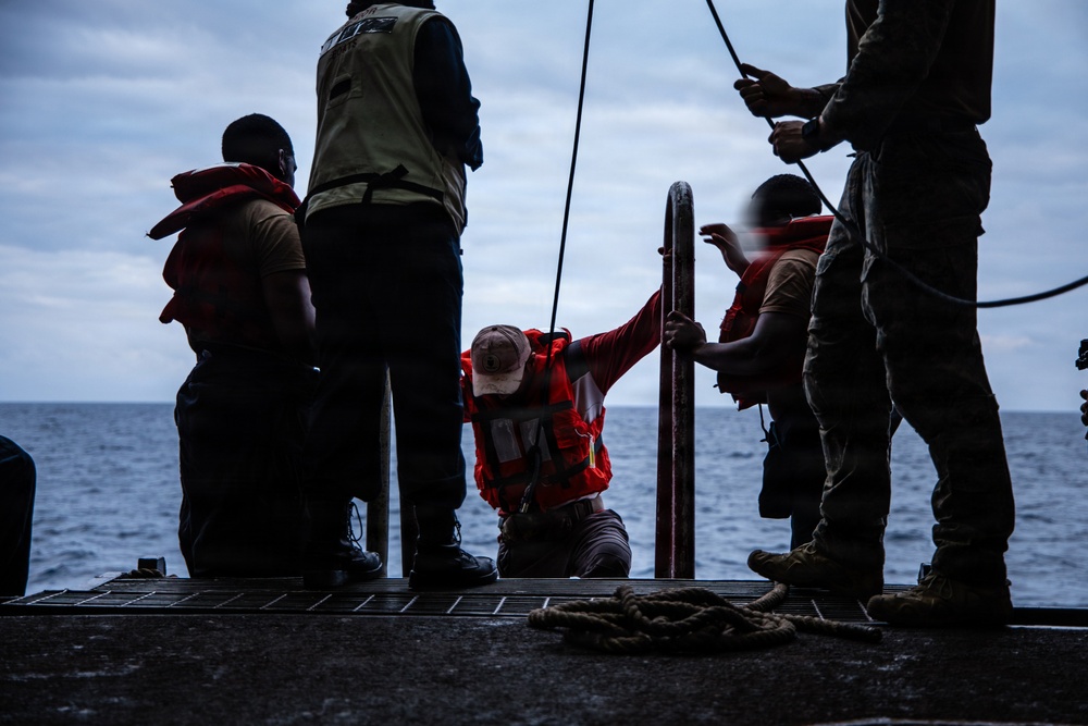 The image shows several individuals on what appears to be the deck of a naval or military vessel at sea. Multiple people are wearing life vests or personal flotation devices, suggesting a maritime safety operation or deck activity. The scene is backlit and silhouetted against an overcast sky and open ocean.