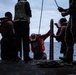 The image shows several individuals on what appears to be the deck of a naval or military vessel at sea. Multiple people are wearing life vests or personal flotation devices, suggesting a maritime safety operation or deck activity. The scene is backlit and silhouetted against an overcast sky and open ocean.