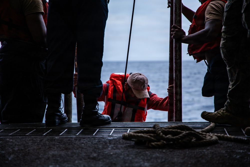A person wearing an orange life vest and a light-colored cap is climbing up onto a vessel deck through an opening or gangway, assisted by crew members standing on either side. The scene appears to take place at sea, with open water visible in the background. This appears to be a maritime rescue or boarding operation.