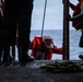 A person wearing an orange life vest and a light-colored cap is climbing up onto a vessel deck through an opening or gangway, assisted by crew members standing on either side. The scene appears to take place at sea, with open water visible in the background. This appears to be a maritime rescue or boarding operation.