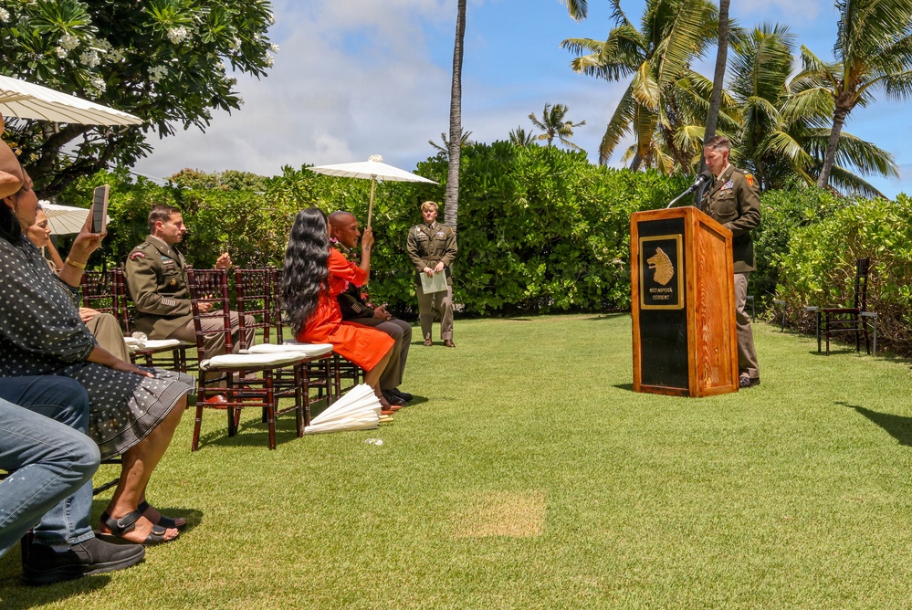 An outdoor military ceremony is taking place on a well-manicured lawn in what appears to be a tropical location, likely Hawaii, based on the palm trees and plumeria. A uniformed military officer is speaking at a wooden podium bearing an emblem, while attendees seated in dark wooden chairs listen. Several military personnel in olive/green uniforms are present alongside civilian guests.