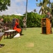 An outdoor military ceremony is taking place on a well-manicured lawn in what appears to be a tropical location, likely Hawaii, based on the palm trees and plumeria. A uniformed military officer is speaking at a wooden podium bearing an emblem, while attendees seated in dark wooden chairs listen. Several military personnel in olive/green uniforms are present alongside civilian guests.