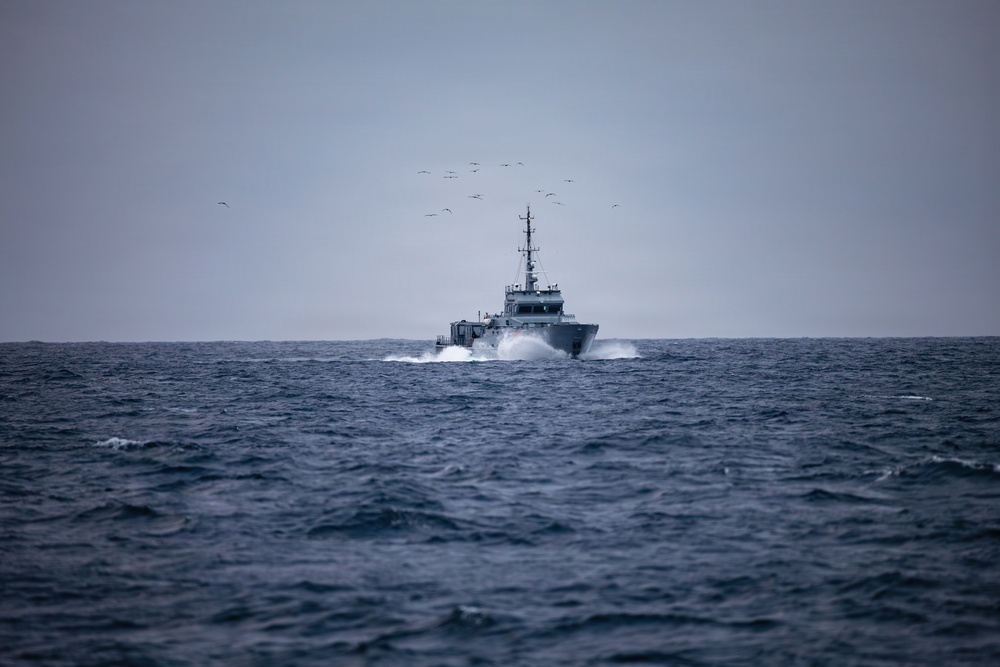 A gray military or coast guard patrol vessel is underway at speed on open, choppy ocean waters under an overcast sky. The vessel is creating a visible bow wave and wake, indicating significant forward momentum. A flock of seabirds is visible in the sky above and behind the vessel.